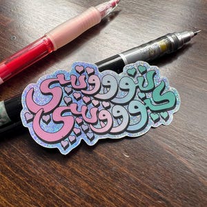 Puede incluir: Una pegatina holográfica con escritura árabe en rosa, azul claro y verde, adornada con pequeñas formas de corazón. La pegatina está colocada sobre una superficie de madera oscura, con un bolígrafo rojo y un bolígrafo negro en el fondo.