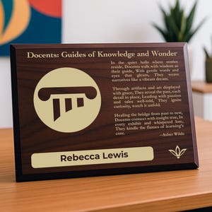May include: A wooden plaque with the text "Docents: Guides of Knowledge and Wonder" and a gold-colored logo. The plaque also includes a name and a poem. The plaque is on a wooden table.