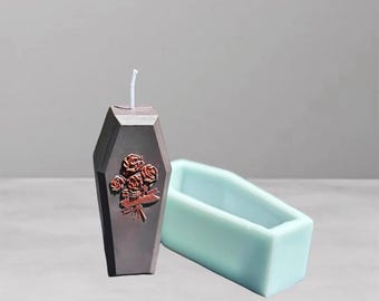Pode incluir: Uma vela cinza escuro em forma de caixão com um buquê de rosas em relevo na frente. Uma forma de caixão azul claro está ao lado da vela. A vela tem um pavio branco.