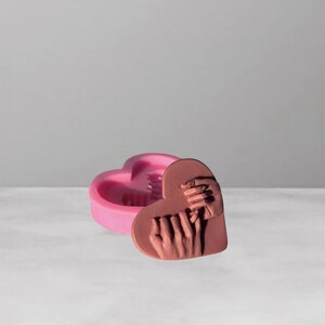Puede incluir: Un molde rosa en forma de corazón con una tapa a juego. La tapa presenta un relieve detallado de dos manos entrelazadas. El molde está diseñado para manualidades, posiblemente para jabón u otros objetos pequeños.