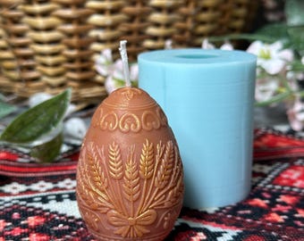 May include: A brown, egg-shaped candle with a wheat design and a blue cylindrical mold. The candle has a wick and is placed on a patterned fabric. A wicker basket is in the background.