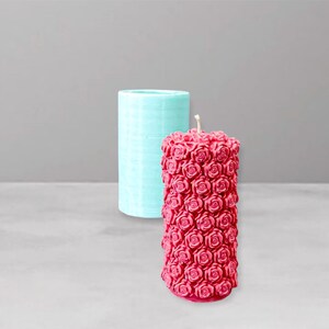 Può includere: Una candela rossa a forma di rosa accanto a uno stampo per candele cilindrico azzurro. La candela è ricoperta da piccole forme di rosa e ha uno stoppino in cima. Lo sfondo è grigio chiaro.