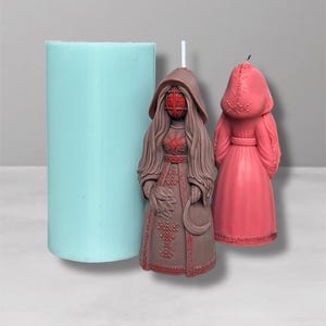 Puede incluir: Un molde cilíndrico de silicona azul claro junto a dos velas decorativas. Una vela es marrón con una máscara roja y detalles intrincados. La otra vela es rosa y presenta un diseño con capucha. Las velas tienen forma de figura.
