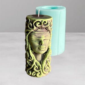 Puede incluir: Una vela verde y marrón con un diseño de rostro con los ojos vendados, junto a un molde cilíndrico azul claro. La vela presenta un rostro esculpido detallado con cabello suelto. La vela es un artículo decorativo único.