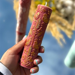 Puede incluir: Una vela de pilar rosa con relieve dorado de p&aacute;jaros y flores. La vela cil&iacute;ndrica tiene una mecha blanca. Otra vela es visible en el fondo, junto con follaje amarillo.