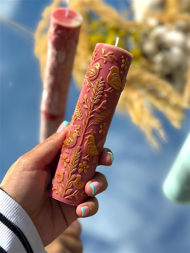 Puede incluir: Una vela de pilar rosa con relieve dorado de p&aacute;jaros y flores. La vela cil&iacute;ndrica tiene una mecha blanca. Otra vela es visible en el fondo, junto con follaje amarillo.