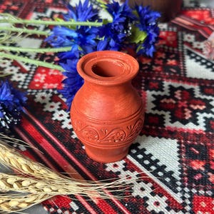 Könnte beinhalten: Eine kleine, terrakottafarbene Vase mit einem detaillierten Blumenmuster in der Mitte. Die Vase steht auf einem gemusterten rot-schwarz-weißen Textil, mit blauen Blumen und Weizenähren in der Nähe. Die Vase ist etwa 10 cm hoch.