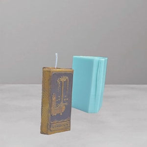 Puede incluir: Una vela rectangular con un diseño dorado y gris, con las palabras "ACE OF SWORDS". Un molde rectangular azul claro está al lado de la vela. La vela tiene una mecha blanca.