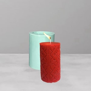 May include: A lit red pillar candle with a detailed geometric pattern, next to a turquoise candle mold. The candle is cylindrical and has a burning wick. The background is a solid gray.