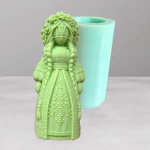 Pode incluir: Um molde de silicone verde claro com um design detalhado de boneca com coroa de flores, tranças e um vestido estampado. Ao lado, um molde cilíndrico, também verde claro. Ideal para projetos de artesanato.
