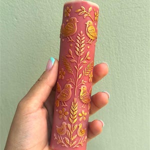 Puede incluir: Una vela cil&iacute;ndrica de color rosa polvoriento con un dise&ntilde;o en relieve dorado de p&aacute;jaros, flores y follaje. La vela tiene una mecha blanca y se sostiene en una mano sobre un fondo verde claro. La vela es un objeto decorativo.