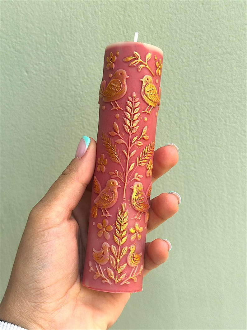 Puede incluir: Una vela cil&iacute;ndrica de color rosa polvoriento con un dise&ntilde;o en relieve dorado de p&aacute;jaros, flores y follaje. La vela tiene una mecha blanca y se sostiene en una mano sobre un fondo verde claro. La vela es un objeto decorativo.