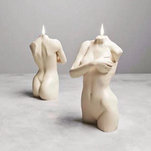 May include: Two ivory-coloured torso candles with lit wicks. One candle is in the foreground, with the other slightly behind it. The candles are sculpted in the shape of a human torso, with detailed anatomical features.