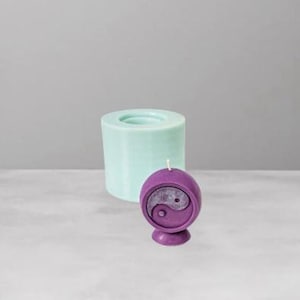 May include: A purple yin-yang candle with a white wick stands next to a light blue cylindrical candle mold. The candle has a rounded shape with a yin-yang symbol in the center. The candle mold is a solid cylinder with a circular indentation.
