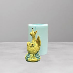 May include: A yellow dragon figurine emerging from a decorative egg, with a light blue cylindrical mold in the background. The dragon has detailed wings and a textured body, and the egg is adorned with a green and yellow base.