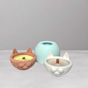 Può includere: Tre portacandele a forma di gatto. Uno è riempito di cera verde chiaro e ha uno stoppino di legno. L'altro è bianco e vuoto. Uno stampo azzurro è dietro di loro. I portacandele sono realizzati in materiale strutturato.