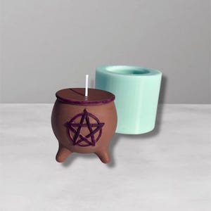 May include: A brown cauldron-shaped candle with a purple pentagram design, a matching lid, and a white wick. A light blue cylindrical mold is next to the candle. The candle has three small feet. The background is a light gray.