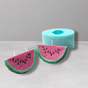 May include: Two watermelon-shaped candles with a pink interior and black seed details, set against a light blue background. The candles have a green rind and white wicks. A light blue cylindrical mould is also present.