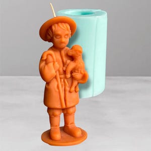 Puede incluir: Vela naranja con forma de pastorcillo sosteniendo un cordero, con un cayado. Un molde de vela cilíndrico azul claro está en el fondo. La vela está sobre una base redonda.