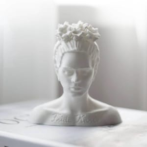 May include: A white bust of Frida Kahlo, featuring a floral crown and the artist's likeness. The sculpture has the name "Frida Kahlo" inscribed on the base. The bust is set against a soft, neutral background.