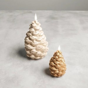 Könnte beinhalten: Zwei kerzenförmige Tannenzapfen. Eine ist groß, cremefarben, die andere kleiner und goldfarben. Beide Kerzen haben einen brennenden Docht. Die Kerzen stehen auf einer hellgrauen Oberfläche.