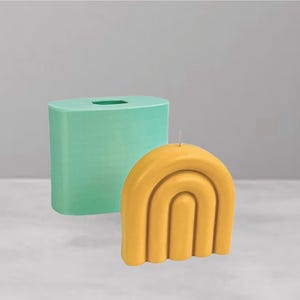May include: A yellow, arch-shaped candle with a wick, next to a light green rectangular candle mould. The candle has a layered arch design. The mould is a solid, rectangular shape with a hole at the top.