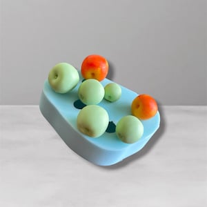 Op de afbeelding: Een lichtblauwe rechthoekige siliconen mal met zes ronde uitsparingen. Twee van de uitsparingen bevatten kleine, ronde, oranje-rode fruitvormige objecten, terwijl de overige vier uitsparingen kleine, lichtgroene fruitvormige objecten bevatten.