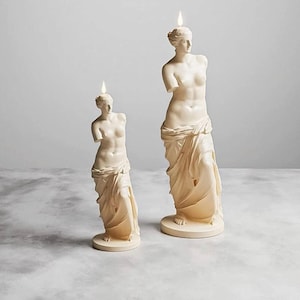 Puede incluir: Dos esculturas de velas de color crema con forma de estatuas clásicas, cada una con una llama en la cabeza. Las estatuas están cubiertas de tela y se apoyan sobre bases circulares. La estatua más grande mide unos 30 cm de altura.