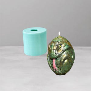 Könnte beinhalten: Eine grün-goldene Schlange-Kerze mit rosa Zunge und gelben Augen. Eine hellblaue zylindrische Kerzenform befindet sich neben der Kerze. Die Kerze hat ein strukturiertes, schuppiges Aussehen.