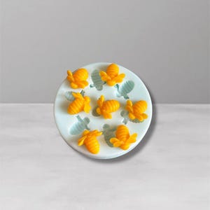 Op de afbeelding: Een witte siliconen mal met zeven gele bijvormige holtes en zes blauwe vleugelindrukken. De mal is ontworpen voor het maken van decoratieve elementen voor bakken of knutselen, met een bijen thema.