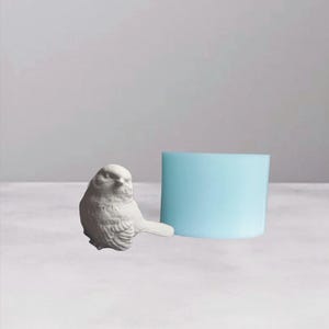 Può includere: Una piccola statuetta di uccello bianco con dettagliate trame di piume è posizionata accanto a un oggetto cilindrico azzurro. L'uccello è posizionato su una superficie bianca contro uno sfondo grigio chiaro. La statuetta ha un aspetto realistico.