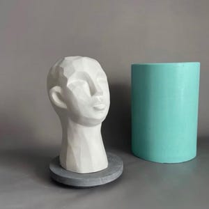 Puede incluir: Una escultura de cabeza blanca de estilo geométrico sobre una base circular gris, junto a un molde cilíndrico turquesa. La cabeza tiene una apariencia lisa y tallada con rasgos faciales definidos. El molde es de color sólido y opaco.