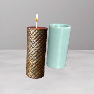 Może przedstawiać: Zapalone świeca z wzorem łuski w odcieniach złota i brązu, obok jasnoniebieskiej cylindrycznej formy. Świeca ma drewnianą górę i jasny płomień.