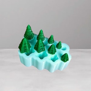 Puede incluir: Un molde de silicona azul claro lleno de formas de árboles de Navidad verdes. El molde tiene un borde ondulado y contiene árboles de varios tamaños. Los árboles son de color verde vibrante y el molde es de un azul pastel suave.