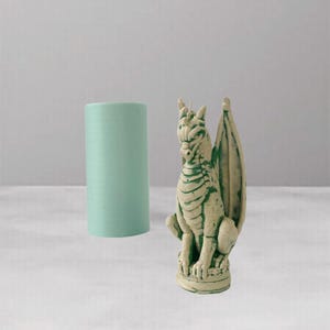 Pode incluir: Uma vela cilíndrica verde claro ao lado de uma estátua de dragão detalhada em branco sujo e verde. O dragão tem asas e está empoleirado em uma base decorativa. A vela e a estátua estão em uma superfície branca.