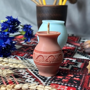 May include: A terracotta-colored candle shaped like a small vase, adorned with floral patterns. The candle has a lit wick and sits on a patterned textile. Other items include blue flowers, wheat stalks, and a brown ceramic pot with yellow candles.