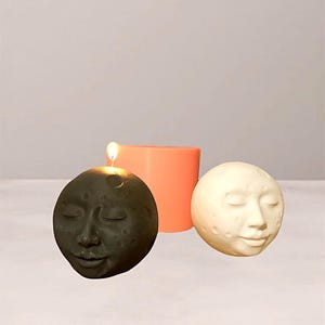 May include: Three moon-shaped candles. One is dark gray with a lit wick, another is off-white, and the third is a peach-colored cylinder. Each candle has a serene face design, perfect for home decor or a unique gift.