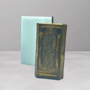 Puede incluir: Una vela rectangular con una imagen detallada de la carta del tarot de la Suma Sacerdotisa. La vela tiene un frente de color bronce con la obra de arte y el texto de la carta. Un molde rectangular azul claro está detrás de la vela.