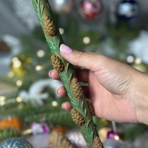 Puede incluir: Una vela verde con detalles de piñas y hojas marrones. La vela es cilíndrica y tiene una estética rústica y natural. El diseño presenta piñas y follaje en relieve, creando un aspecto festivo y de temporada. La vela está sostenida en una mano.
