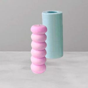 May include: A pink, stacked candle with a cylindrical, light blue candle mold. The candle is made of six rounded sections. The mold is a solid cylinder with a hole at the top. The background is a light gray.