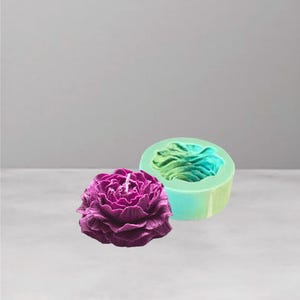 May include: A purple peony-shaped candle with a white wick beside a teal silicone mould. The candle has detailed petal textures. The mould is round with a flower design. The background is a light grey.