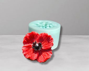 May include: A vibrant red poppy flower with a black center rests beside a light blue cylindrical mold. The poppy has detailed petals and a realistic appearance, suggesting a craft or art project. The background is a soft gray.