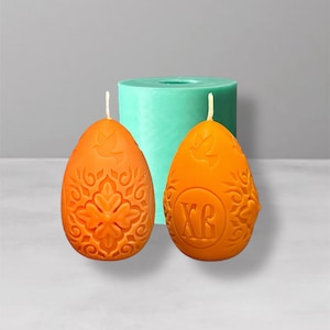 Puede incluir: Dos velas naranjas en forma de huevo con intrincados diseños florales y de palomas. Una vela tiene las letras "XR". Un molde cilíndrico turquesa está detrás de las velas. Las velas tienen mechas blancas.