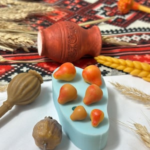 Peut inclure: Un moule en silicone bleu clair avec six cavités en forme de poire, entouré de tiges de blé séchées, deux capsules de pavot brunes, un petit pot en terre cuite et un épi de blé en cire jaune sur un tissu à motifs rouges et blancs.