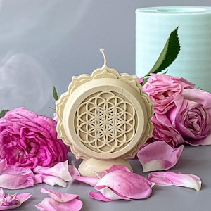 May include: A beige candle with a geometric design, surrounded by pink roses and petals. A light blue cylindrical object is in the background. The candle has a wick and is placed on a gray surface.