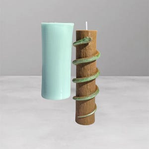 Op de afbeelding: Twee pilaar kaarsen. Eén is lichtblauw, cilindrisch van vorm. De andere is bruin met een groene en gouden slang eromheen gewikkeld. De bruine kaars heeft een witte lont. De kaarsen staan op een lichtgrijze ondergrond.