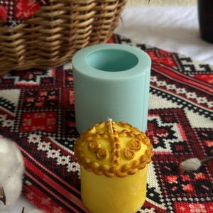 Op de afbeelding: Een gele kaars met een decoratieve bovenkant, naast een turquoise siliconen mal. De kaars heeft een gevlochten ontwerp rond de bovenkant en een lont. De achtergrond omvat een gevlochten mand en een patroontextiel.