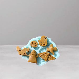 May include: A light blue silicone mold with various Halloween-themed shapes. The shapes include a pumpkin, ghost, bat, and cat. The shapes are a light brown color and are arranged on the mold.