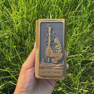 Puede incluir: Una vela rectangular con un marco dorado y un centro gris oscuro, con un dise&ntilde;o en relieve de una vara, hojas y nubes. El texto "ACE of WANDS" est&aacute; en la parte inferior. La vela se sostiene contra un fondo de hierba verde.