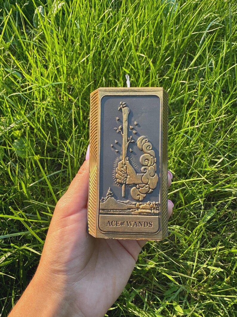 Puede incluir: Una vela rectangular con un marco dorado y un centro gris oscuro, con un dise&ntilde;o en relieve de una vara, hojas y nubes. El texto "ACE of WANDS" est&aacute; en la parte inferior. La vela se sostiene contra un fondo de hierba verde.